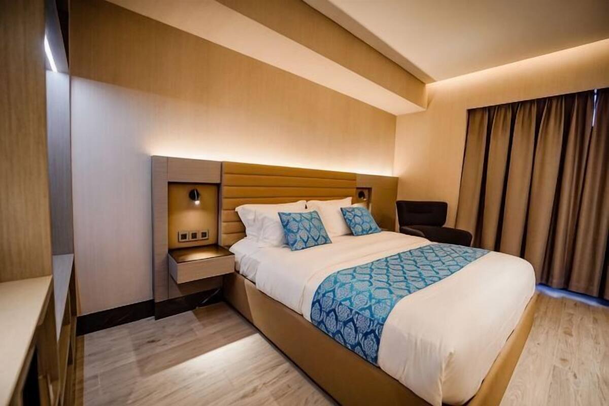 Ein Hotelzimmer mit einem großen Bett, Nachttischen, einem Nachtschränkchen, einem Kopfteil und einem Fenster mit Vorhängen.