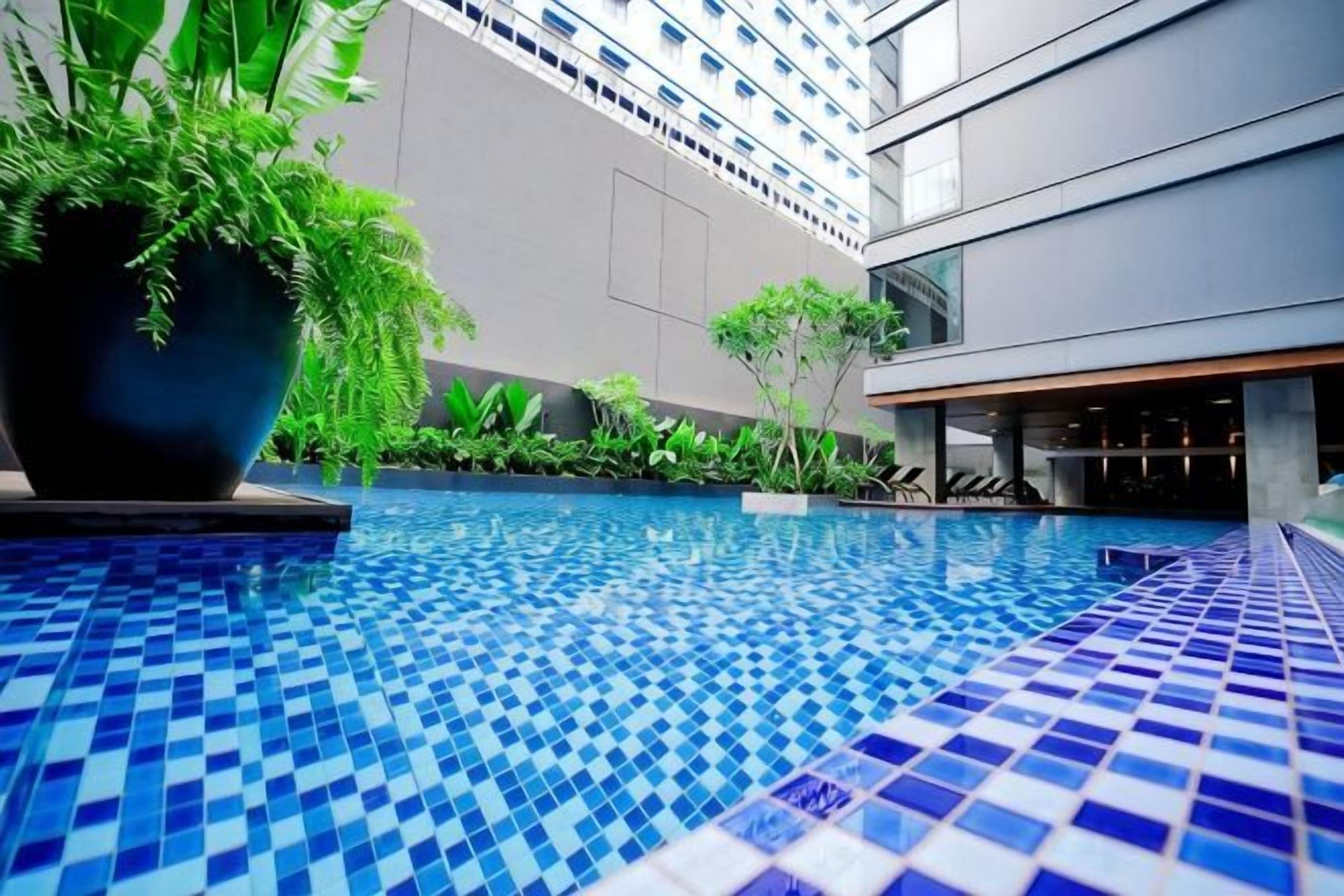 A swimming pool with a tiled floor, surrounded by greenery and a modern building in the background.