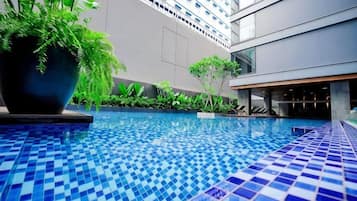 A swimming pool with a tiled floor, surrounded by greenery and a modern building in the background.