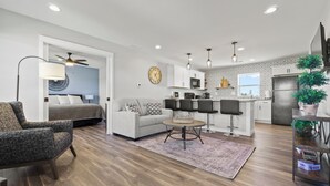 A modern living room with a sofa, armchairs, and a coffee table. There is a kitchen area with cabinets, a refrigerator, and a dining area with bar stools. - Country Club Cottages - 1703 (Washington)