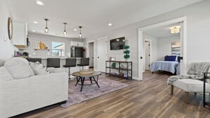A modern living room with a sofa, a coffee table, and a TV. There is a kitchen area with a fridge and a dining table. The room has wooden flooring and a large window. - Country Club Cottages - 1703 (Washington)