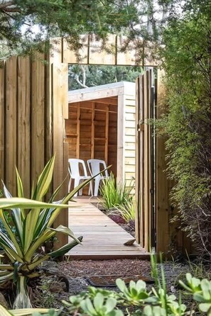 A wooden deck leading to a room with a sloped roof, surrounded by greenery and white plastic chairs. - Orchard Cottage—Romantic Bushland Wellness Retreat (Yallingup Siding)