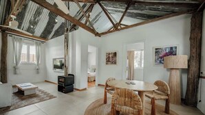 A spacious living room with a high ceiling, a dining area with a round table and wicker chairs, a fireplace, and a television. - Orchard Cottage—Romantic Bushland Wellness Retreat (Yallingup Siding)