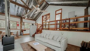 A spacious room with a high ceiling, a sofa, a coffee table, a bookshelf, and a staircase. - Orchard Cottage—Romantic Bushland Wellness Retreat (Yallingup Siding)