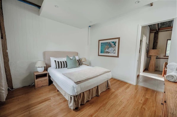 Un dormitorio con cama, mesitas de noche, lámpara, un cuadro y suelo de madera.