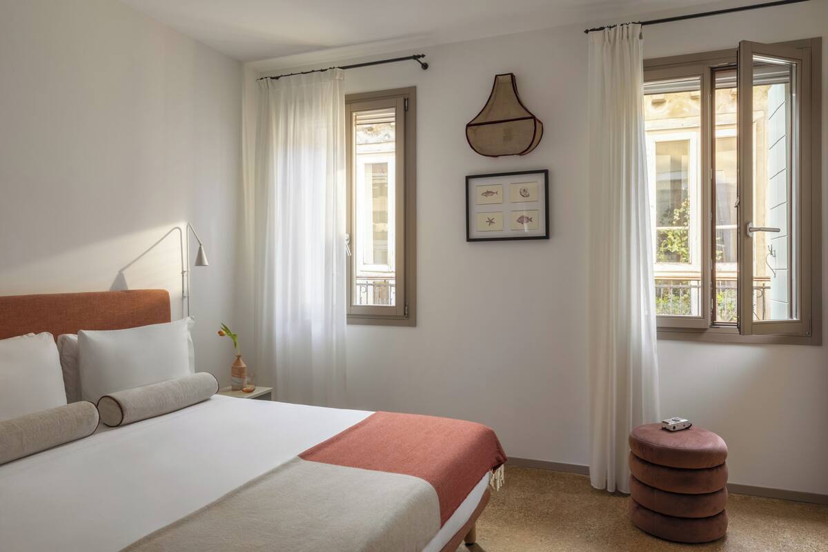 A bedroom with a bed, a nightstand, a lamp, a window with curtains, and a wall-mounted picture.