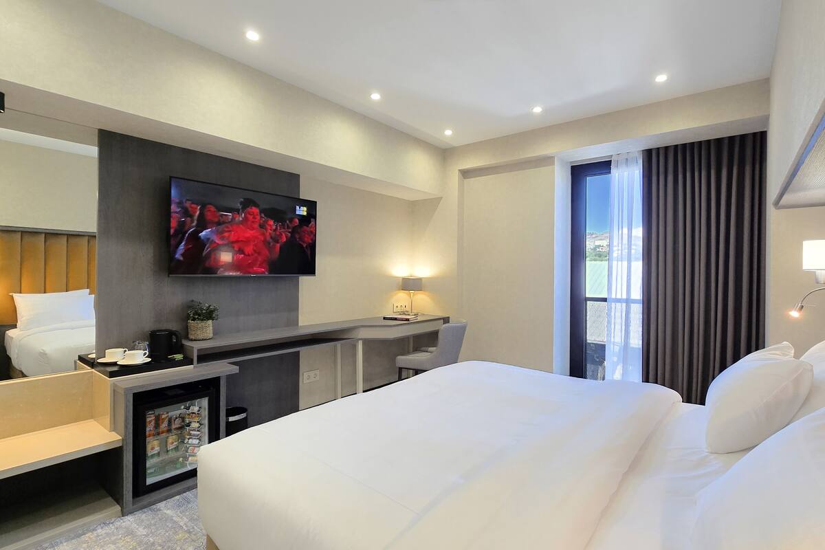 Ein Hotelzimmer mit einem großen Bett, einem Schreibtisch mit Stuhl, einem an der Wand montierten Fernseher und einem Fenster mit Vorhängen.