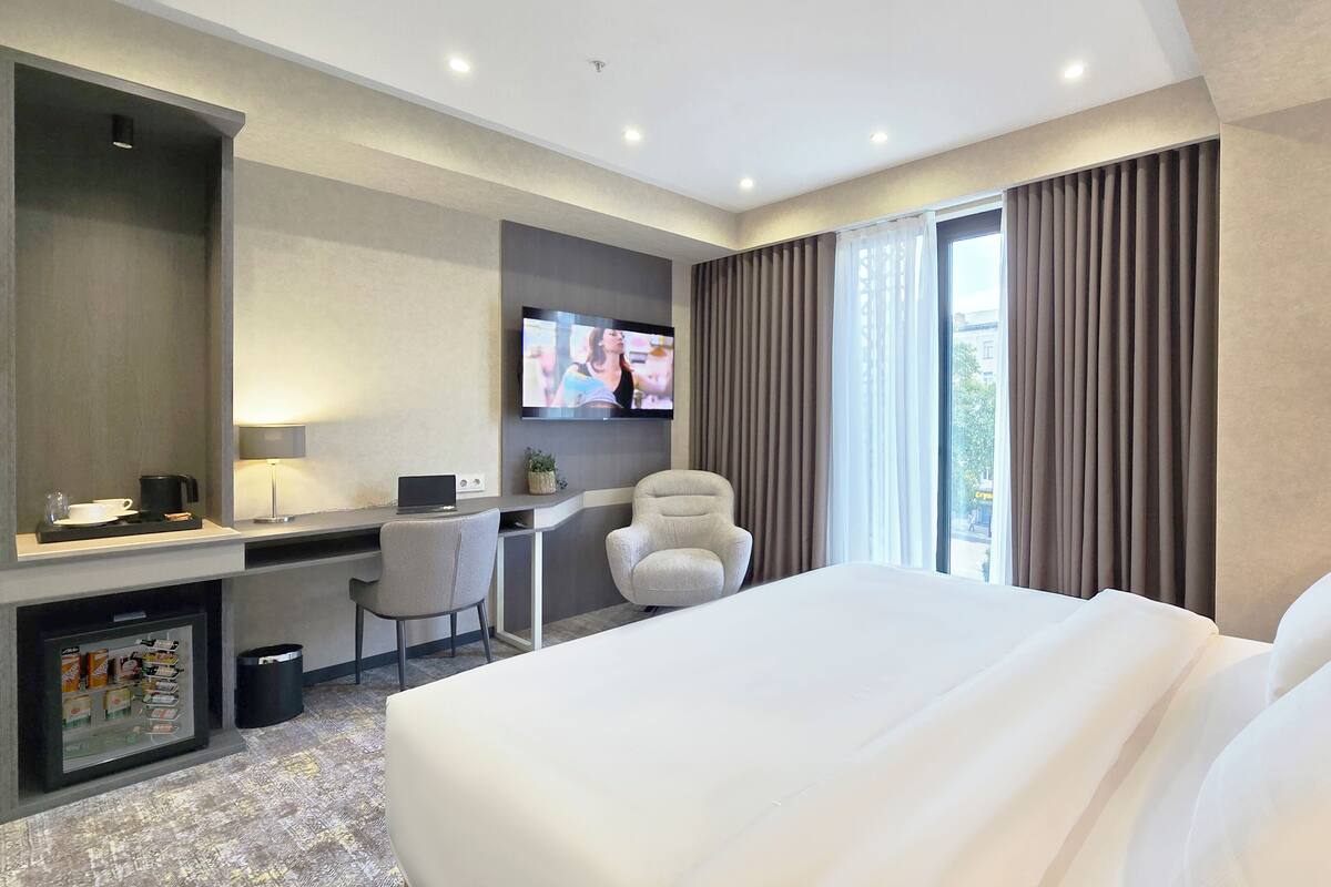 Ein Hotelzimmer mit einem großen Bett, einem Schreibtisch mit einem Laptop, einem Sessel, einem Fernseher und einem Fenster mit Vorhängen.