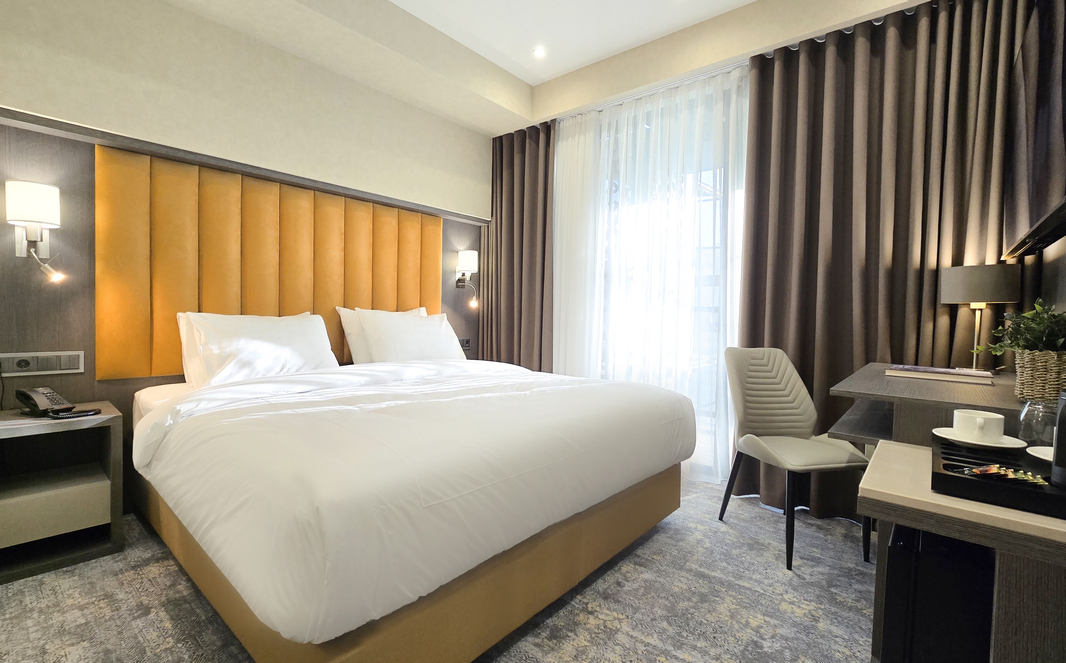 A hotel room with a large bed, a desk with a lamp, a chair, and a view of the outdoors.