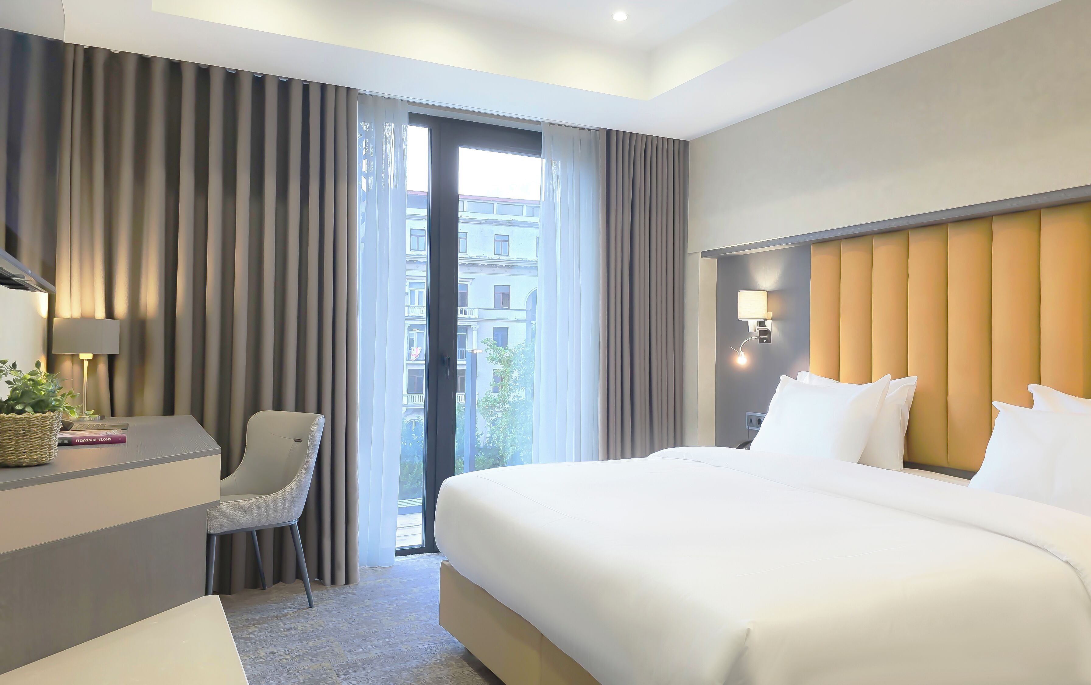 Ein Hotelzimmer mit Bett, Schreibtisch mit Lampe, Stuhl und Blick auf Gebäude durch das Fenster.