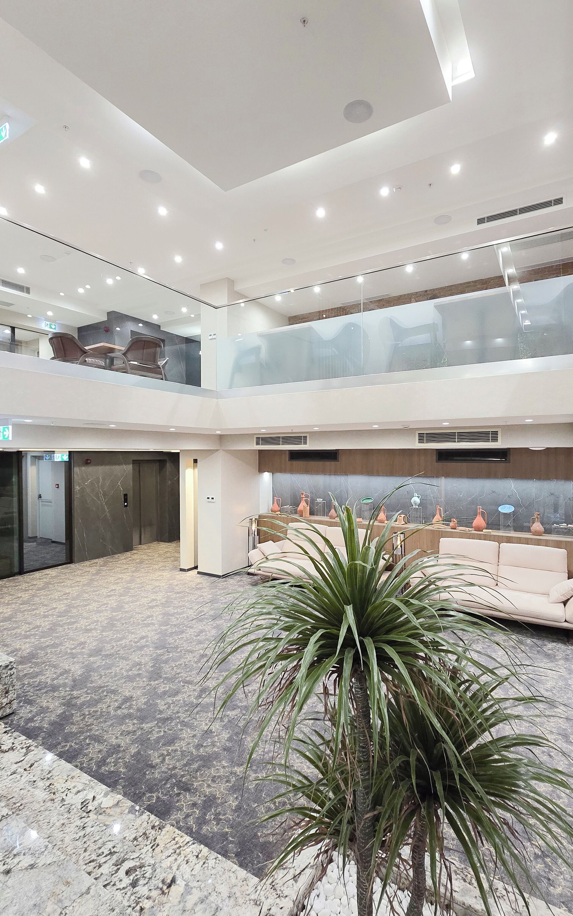 A spacious lobby with a marble floor, a central plant, seating area, and a mezzanine level with a view of the lobby.