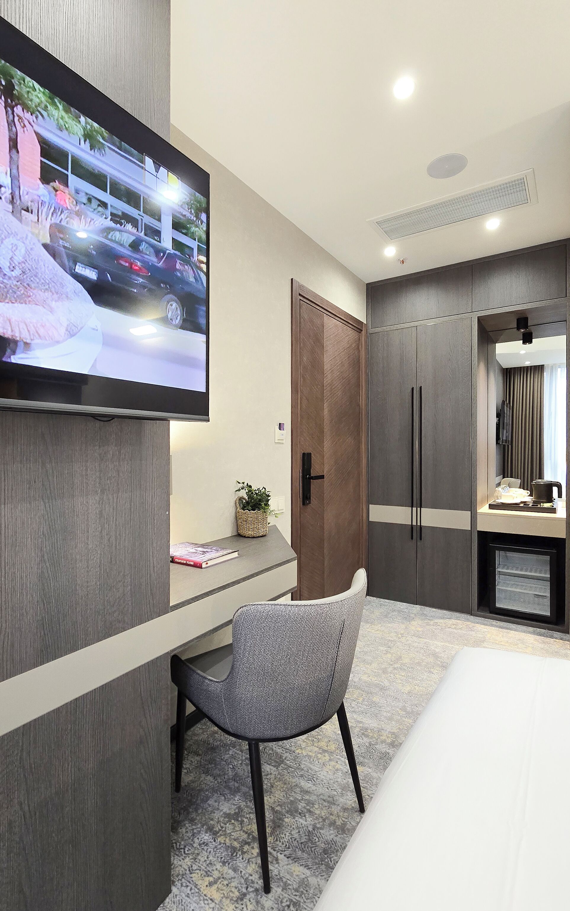 A modern hotel room with a large flat-screen TV mounted on the wall, a desk with a chair, a wardrobe, and a bed.