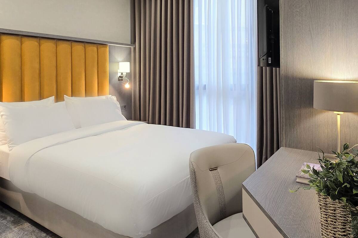 Ein modernes Hotelzimmer mit einem großen Bett, einem Schreibtisch mit Stuhl, einem Fernseher und einem Fenster mit Vorhängen.