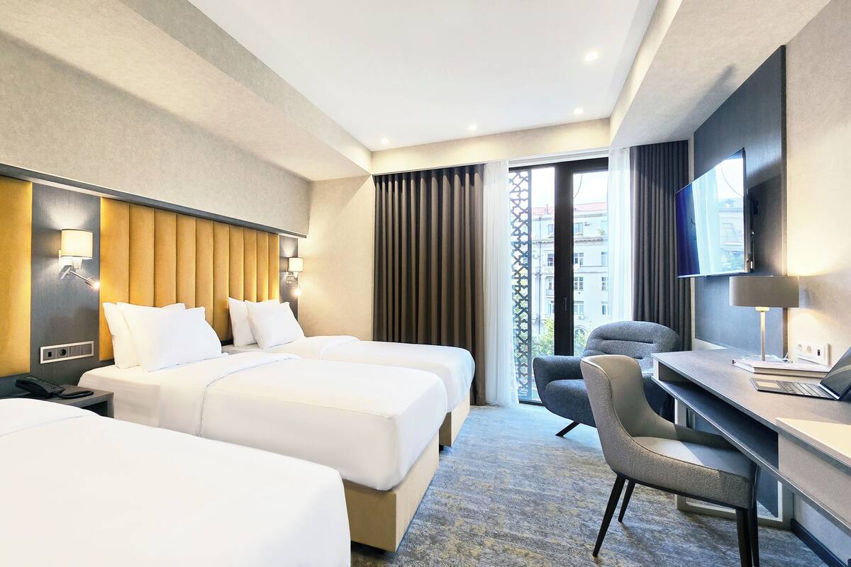 Ein Hotelzimmer mit zwei Betten, einem Schreibtisch mit Stuhl, einem Fernseher und einem Fenster mit Vorhängen.