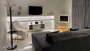 A modern hotel room with a sofa, a small round table, a white chair, a TV, a fireplace, and a coat rack. - Angel's Apartment Volos (Volos)
