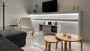 A modern living room with a grey sofa, two wooden tables, a white chair, a flat-screen TV mounted on a white wall, and a coat rack with a black umbrella. - Angel's Apartment Volos (Volos)