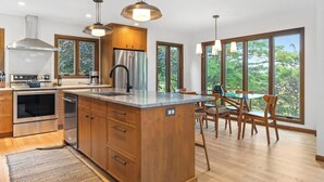 A modern kitchen with wooden cabinets, stainless steel appliances, and large windows offering a view of the outdoors. - The Hideout: Luxe Escape w/ Sauna & Hot Tub | Birchwood Golf & CC (Harbor Springs)