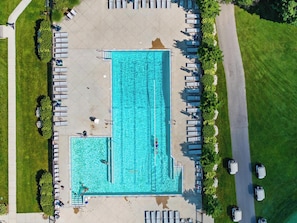 Piscina retangular com deck ao redor, cadeiras de descanso e área de estacionamento.