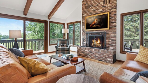 A cozy living room with a fireplace, leather sofas, and large windows offering a view of the outdoors. - The Hideout: Luxe Escape w/ Sauna & Hot Tub | Birchwood Golf & CC (Harbor Springs)