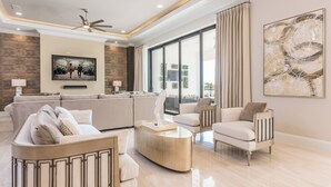 A spacious living room with a large sofa, a coffee table, and a seating area with armchairs. There is a large window with curtains, a ceiling fan, and a painting on the wall. - The Superhero Haven-TT276 (Kissimmee)