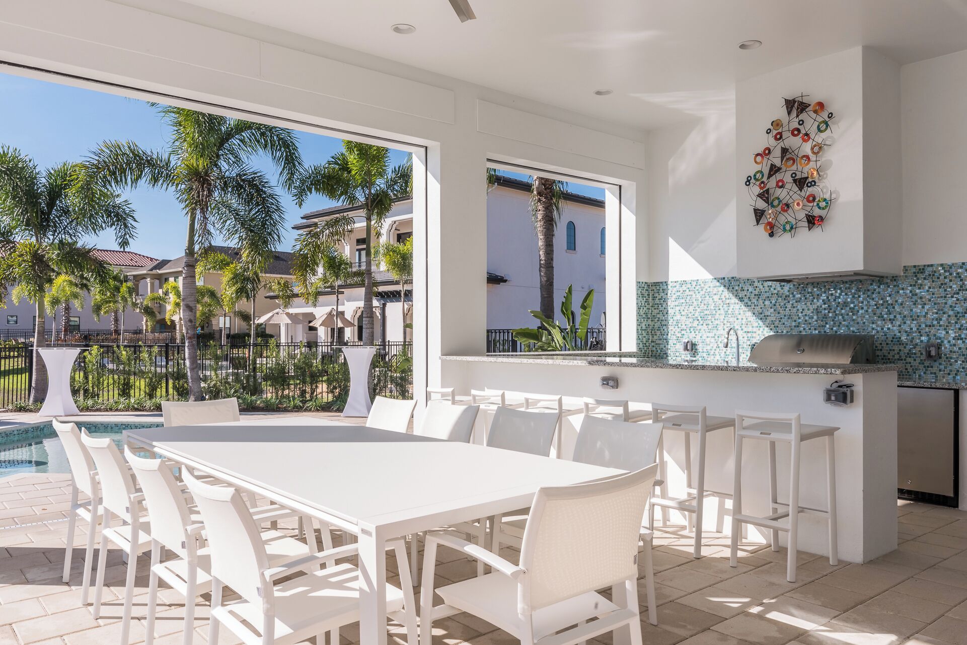 A modern outdoor kitchen with a dining area, a pool, and a view of palm trees and buildings.