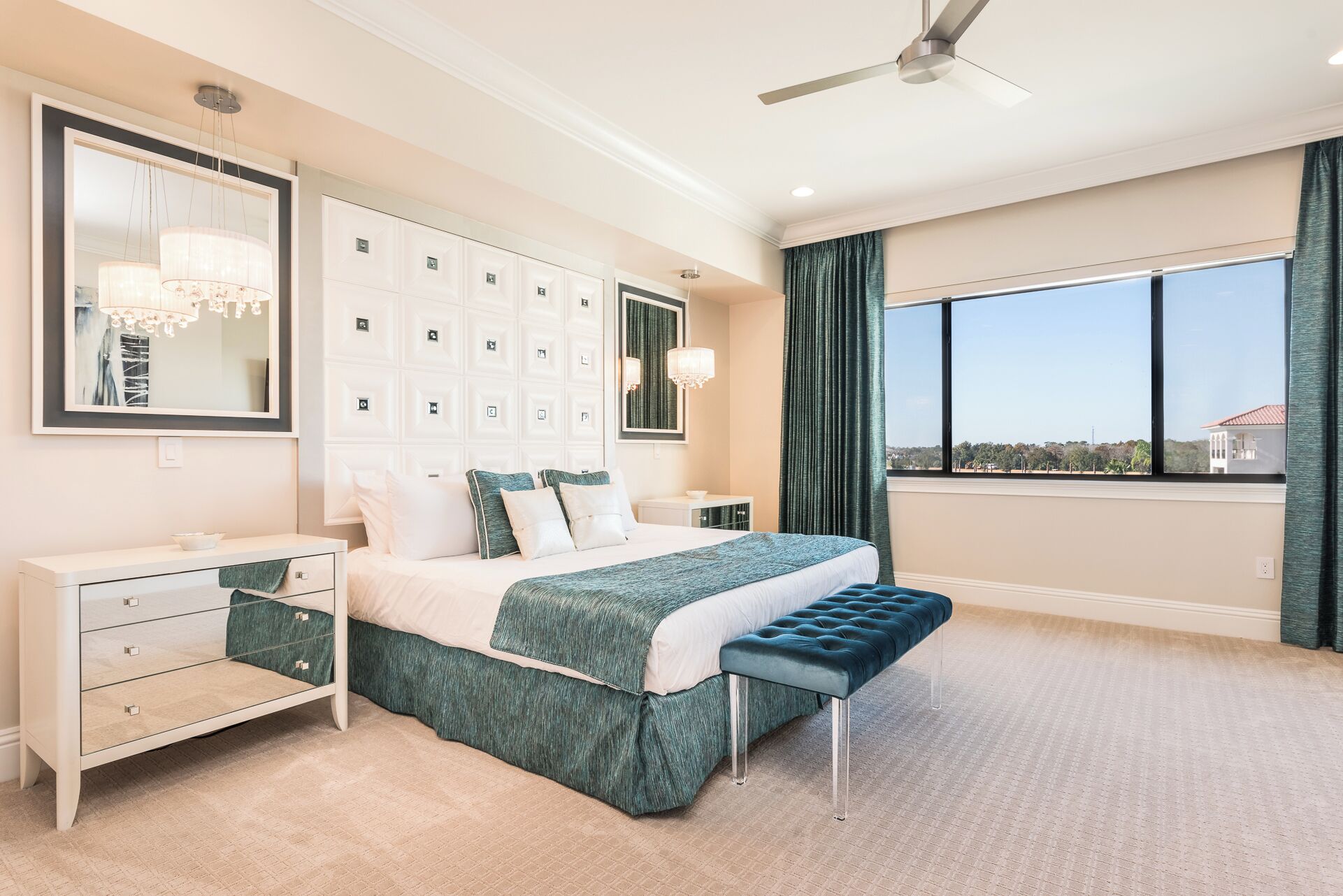 A modern bedroom with a large bed, a vanity with drawers, a bench, a ceiling fan, and a large window with a view.