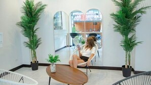 Un espace intérieur moderne où l’on voit une personne assise sur une chaise, deux plantes en pot et une petite table sur laquelle est posé un vase.