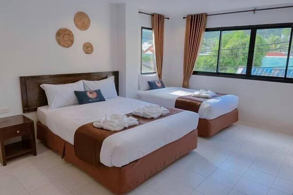 A hotel room with two beds, a nightstand, a window with curtains, and a view of greenery outside.