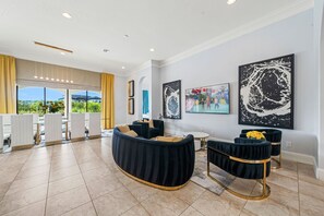 A modern living room with a curved black sofa, a round coffee table, and a dining area with white chairs and a glass table. There are two black and gold side tables with yellow flowers. The room has large windows with a view of greenery and a building. - The Family Fun House-TT350 (Kissimmee)