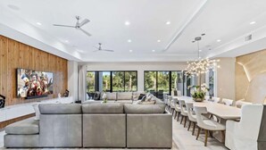 Un salon moderne avec un canapé d’angle, une table à manger et des chaises. On y trouve des ventilateurs de plafond, de grandes fenêtres et un lustre.