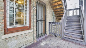 Eine hölzerne Treppe führt zu einer Tür mit einem Metallgitter, einem Fenster mit Metallstreben und einer Ziegelwand.