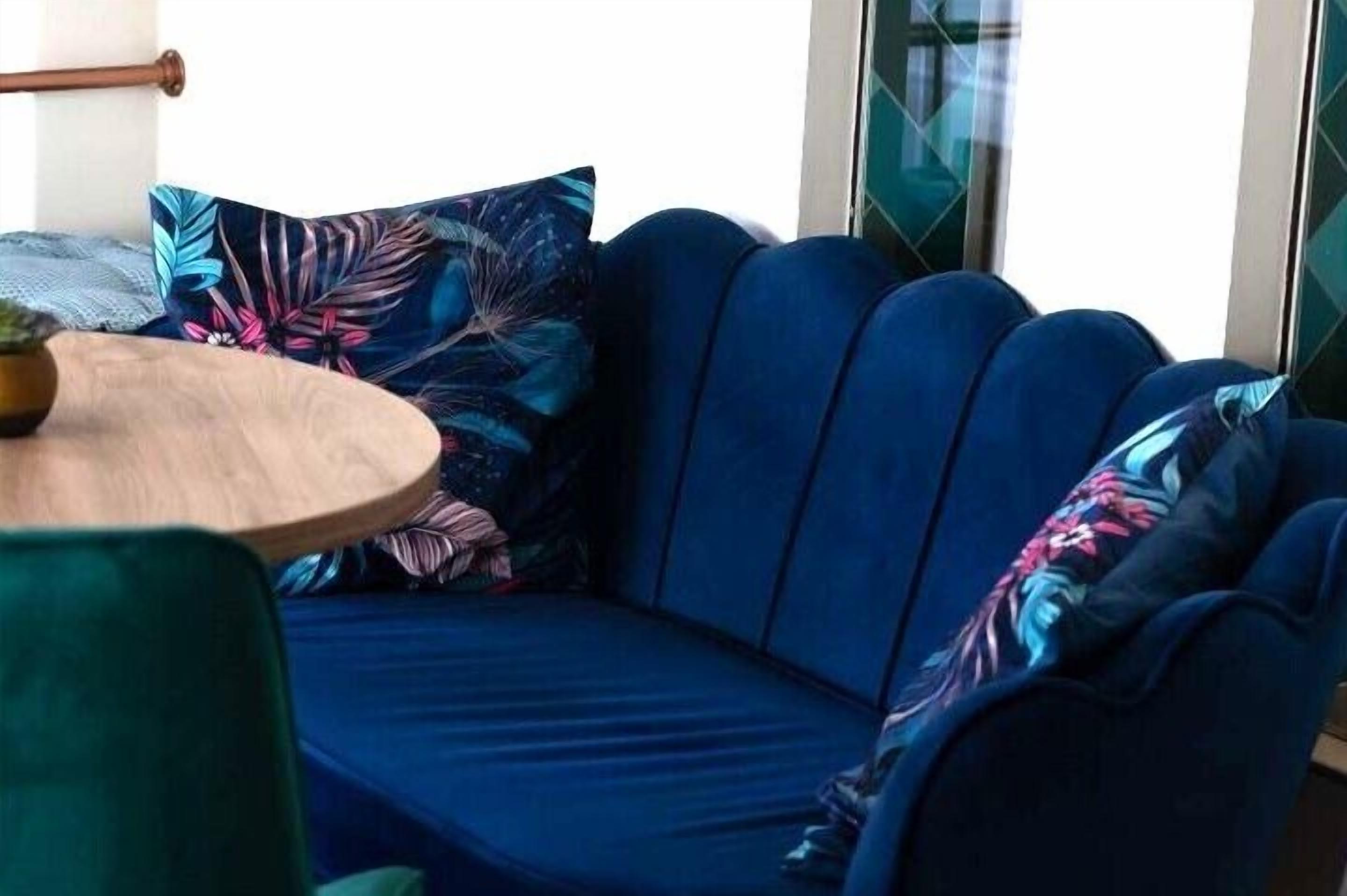 A blue sofa with a floral pillow, a round wooden table, and a green chair in a modern room with glass walls.
