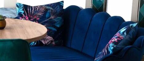A blue sofa with a floral pillow, a round wooden table, and a green chair in a modern room with glass walls.