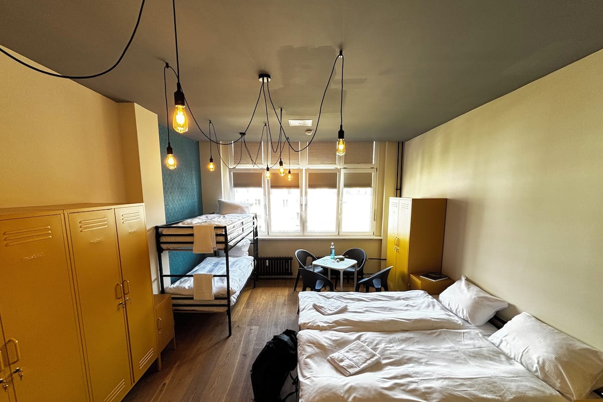 A dormitory room with bunk beds, a desk, and a chair. There are lockers on one side and a window providing natural light.