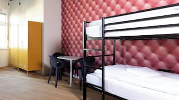 Una cama litera con cabecera tapizada roja, un escritorio, dos sillas y un armario amarillo en una habitación con suelo de madera.