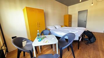 A hotel room with a bed, a desk with chairs, a wardrobe, and a table with a bottle and cards.