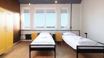 A dormitory room with two single beds, wooden flooring, a radiator, and a window with blinds.