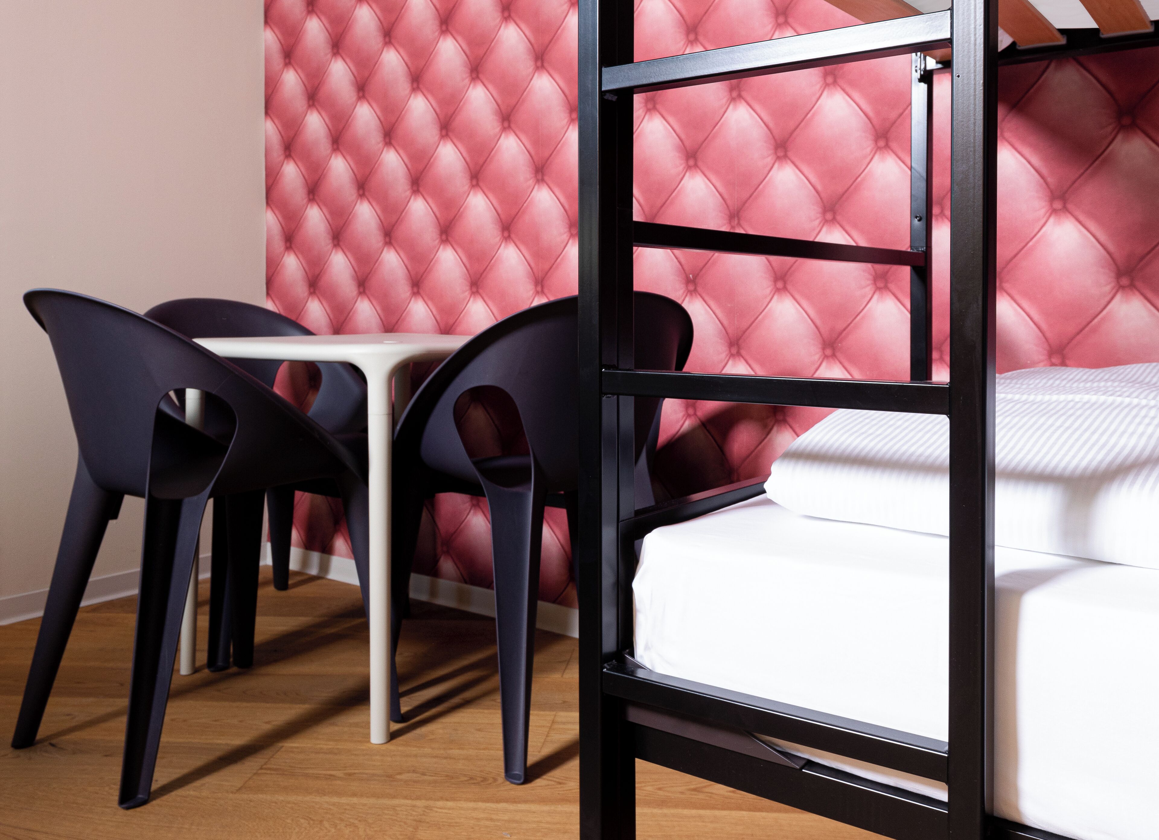 A bunk bed with a white mattress and a red tufted headboard. There is a small white table and two black chairs in front of the bed.