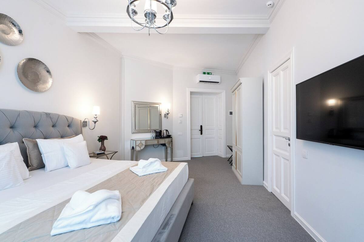 A hotel room with a bed, a vanity with a mirror, a flat-screen TV mounted on the wall, and a door leading to another room.