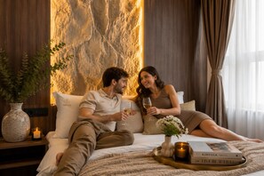 A couple sitting on a bed with a textured wall behind them, a bedside table with a candle, and a tray with books and a glass. - Highline Downtown Aparthotel (Bucharest)