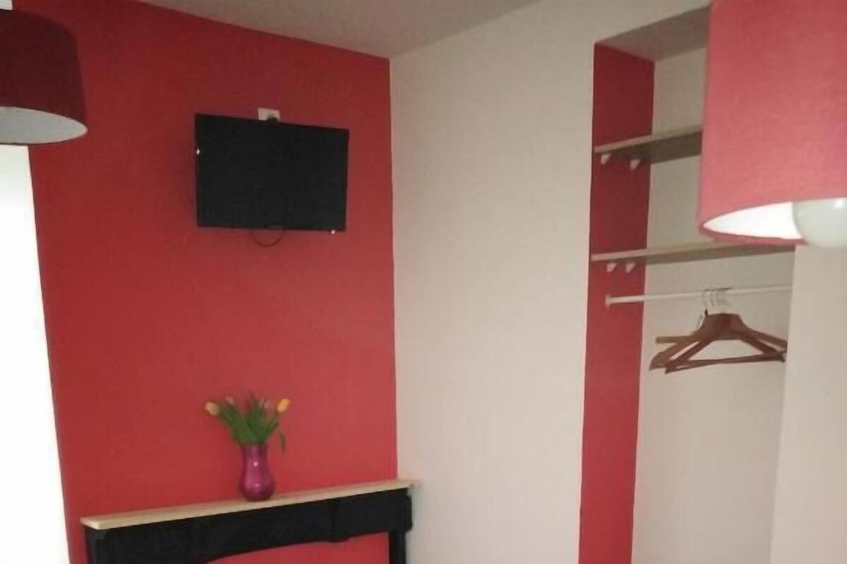 Ein Bett mit weißer und roter Bettwäsche, ein kleiner Tisch mit Blumen in einer Vase und ein an der Wand befestigter Fernseher.