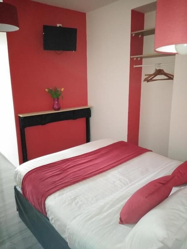 Uma cama com roupa de cama branca e vermelha, uma mesinha com um vaso de flores e uma televisão presa na parede.