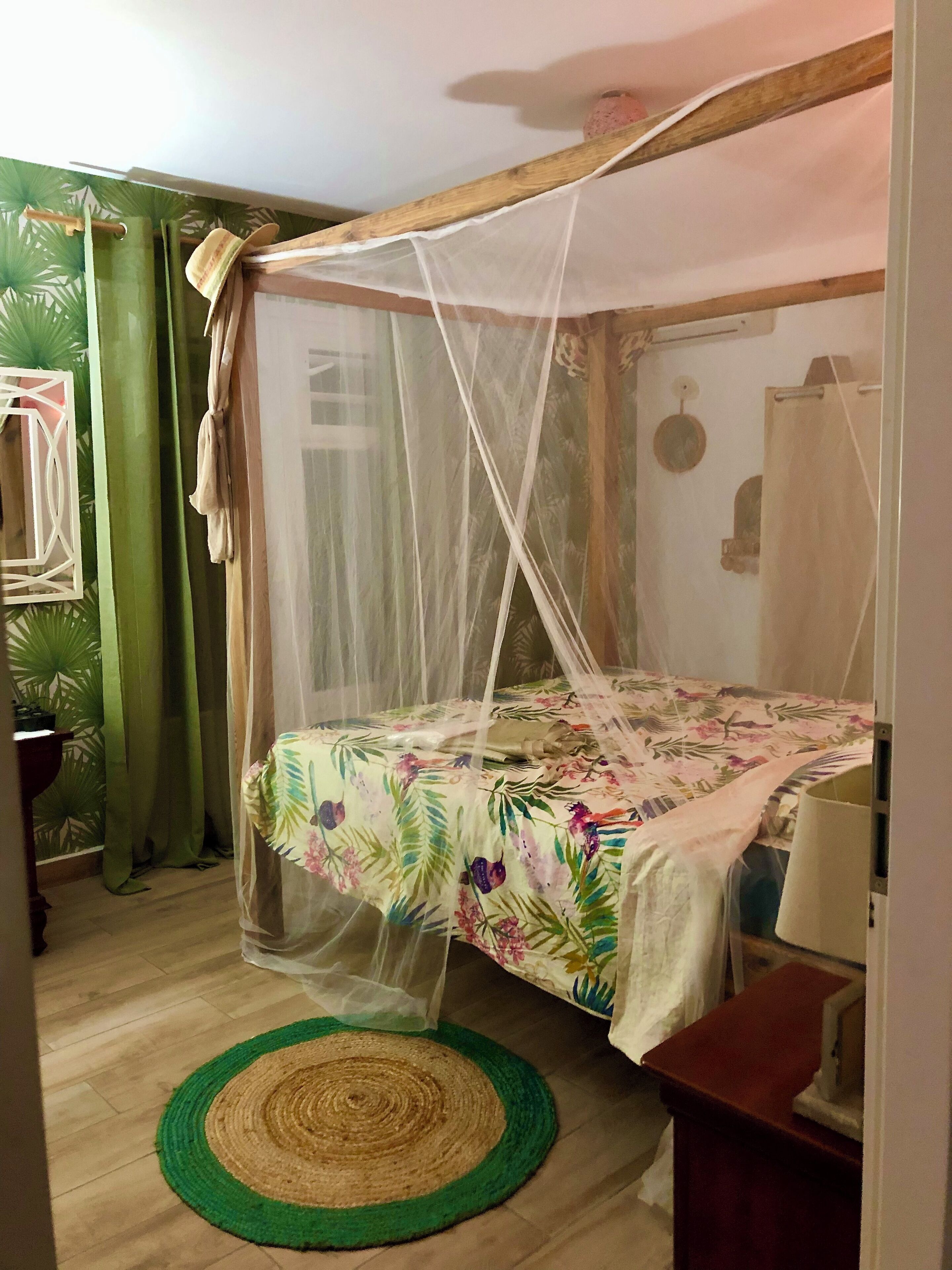 Una habitación con una cama bajo una mosquitera, un tapete tejido verde y marrón, y un cabecero de madera.