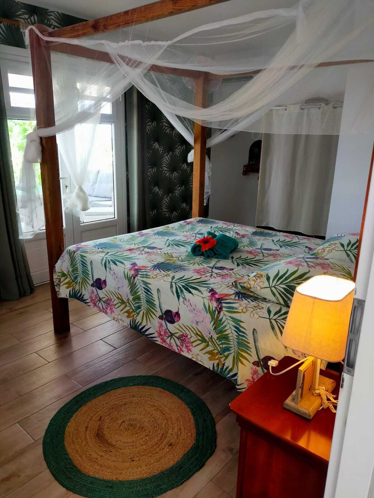 A bedroom with a bed under a canopy, a bedside table with a lamp, and a window with curtains.