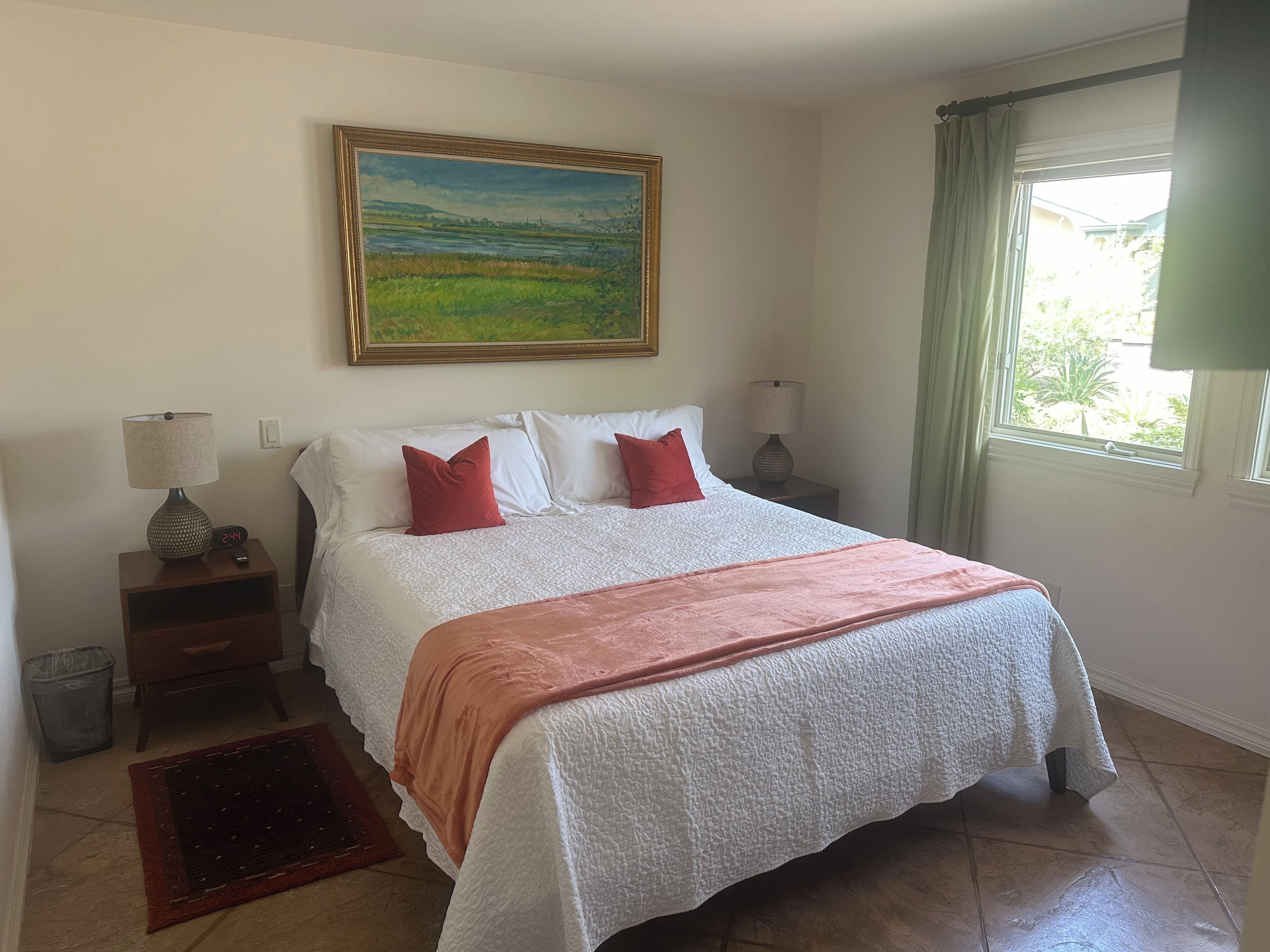 A bedroom with a large bed, two bedside tables with lamps, a painting on the wall, and a window with curtains.