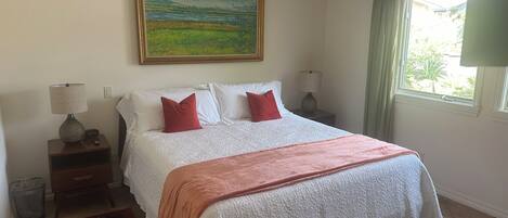 A bedroom with a large bed, two bedside tables with lamps, a painting on the wall, and a window with curtains.