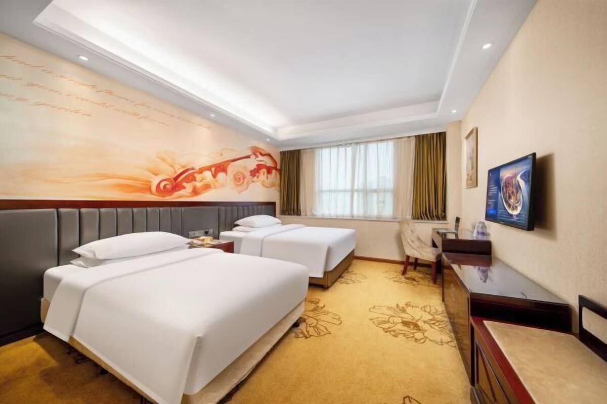 A hotel room with two beds, a desk, a television, and a large window with curtains.