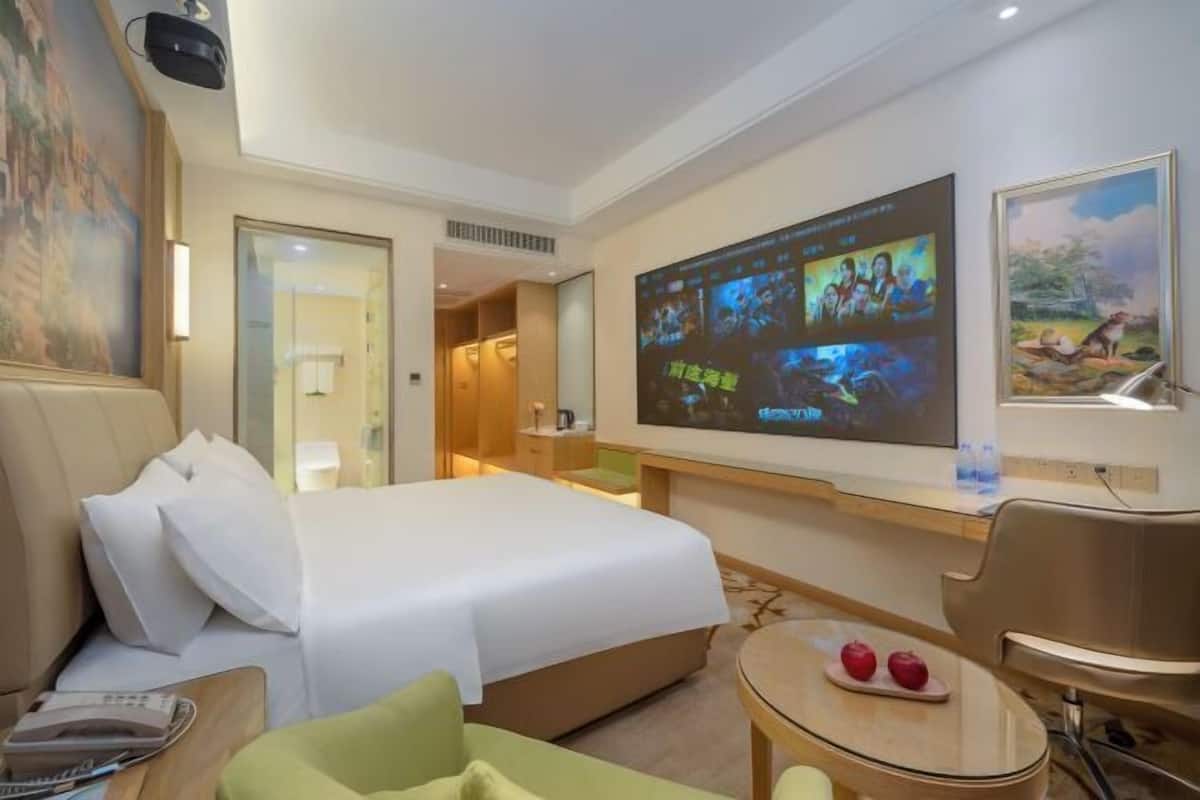 A hotel room with a bed, a desk, a chair, a TV screen, and a painting on the wall.