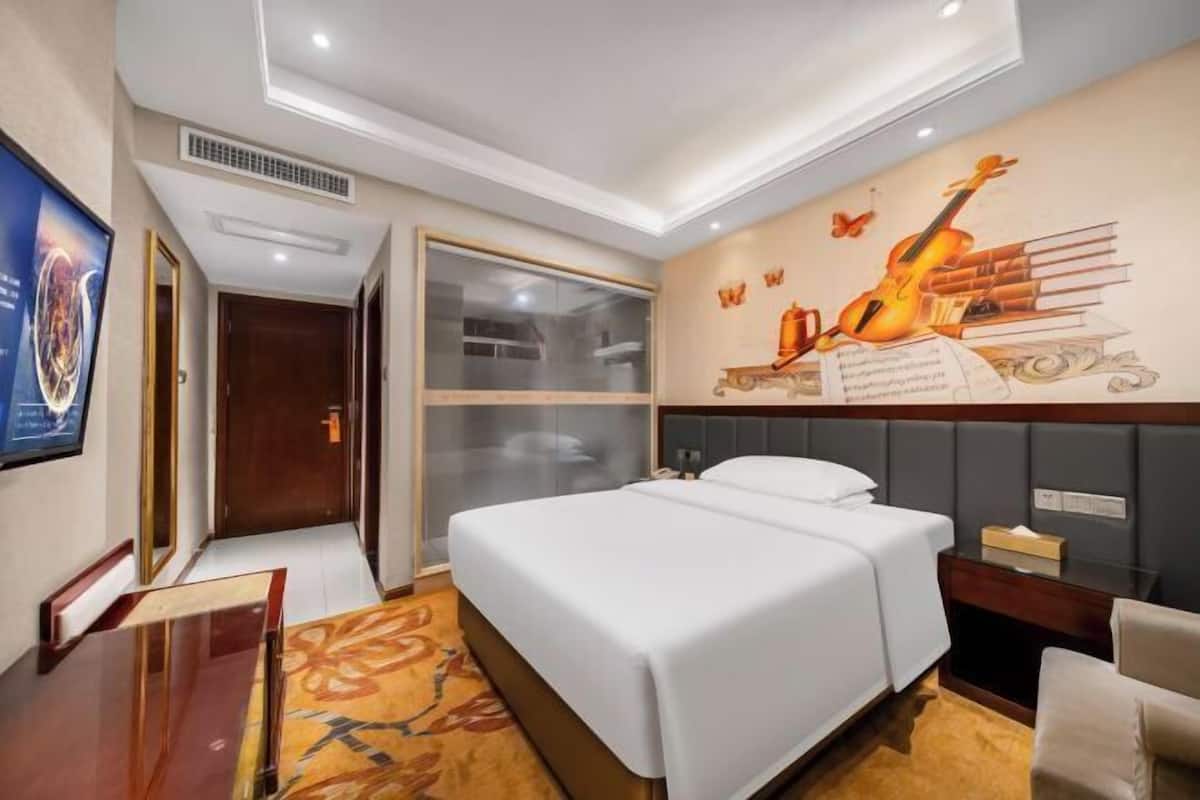 A hotel room with a bed, a desk, a chair, a wall mural, and a large window.