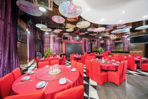 Een balzaal met ronde tafels, rode tafelkleden en paarse gordijnen.